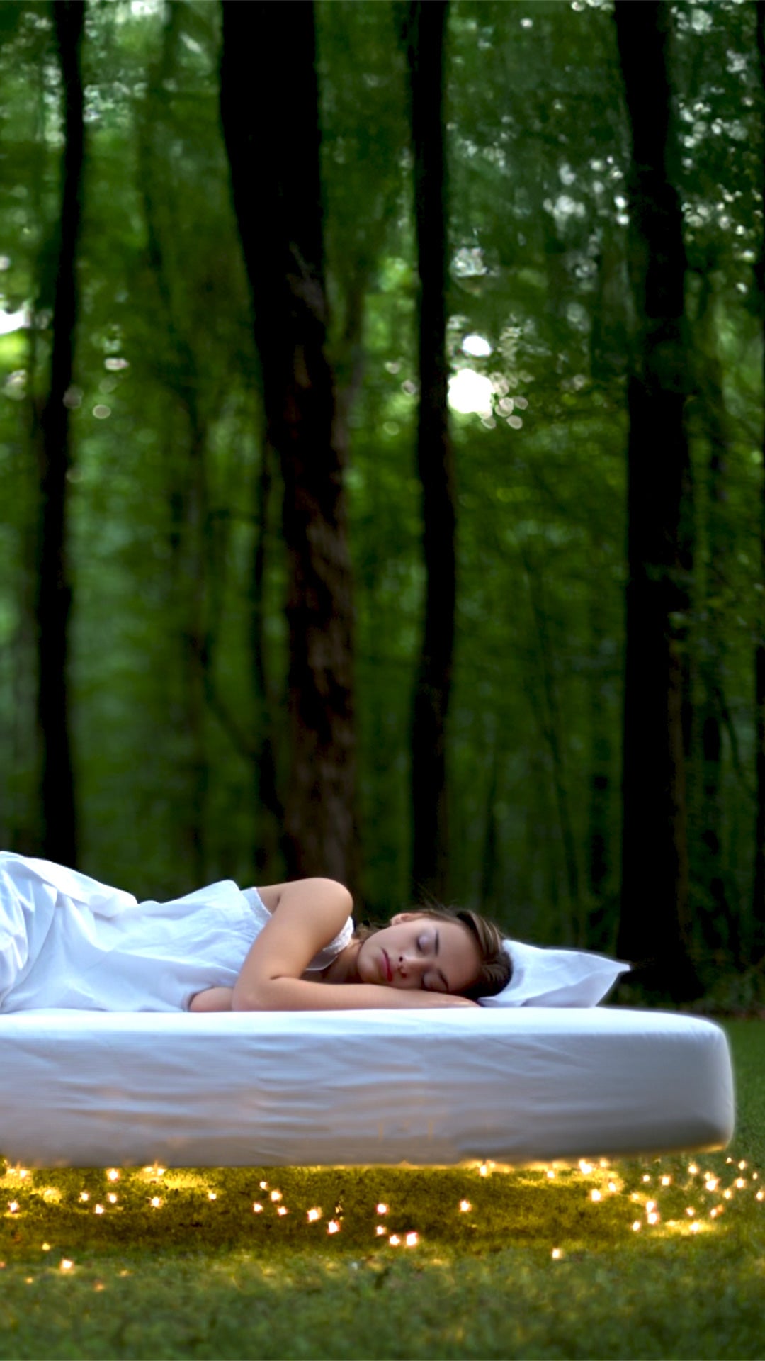 A woman sleeping soundly on a white Elvar® mattress floating above a grassy lawn with warm fairy lights and a dense forest background.