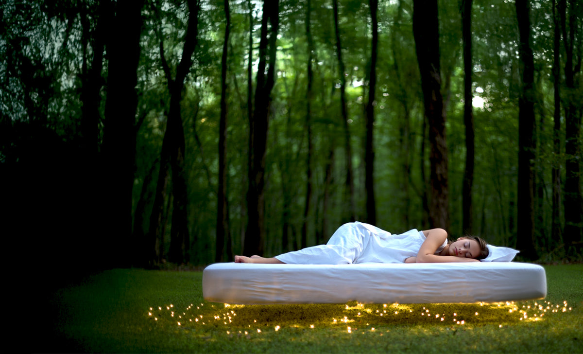 A person lies on a white Elva® mattress floating above a grassy lawn with a forest backdrop, surrounded by small warm lights.