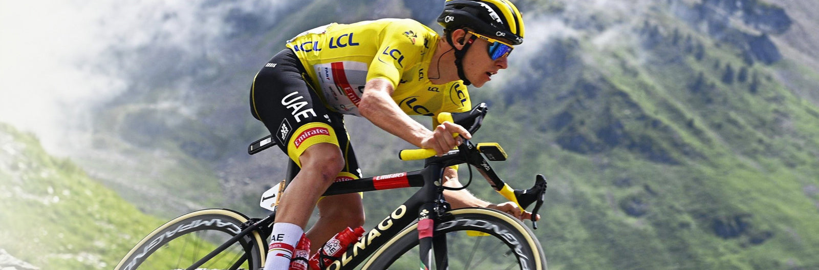 A UAE cyclist in the Tour de France in a yellow jersey rides a black road bike through a mountainous green landscape. The bike has visible Colnago branding on the frame and wheels.