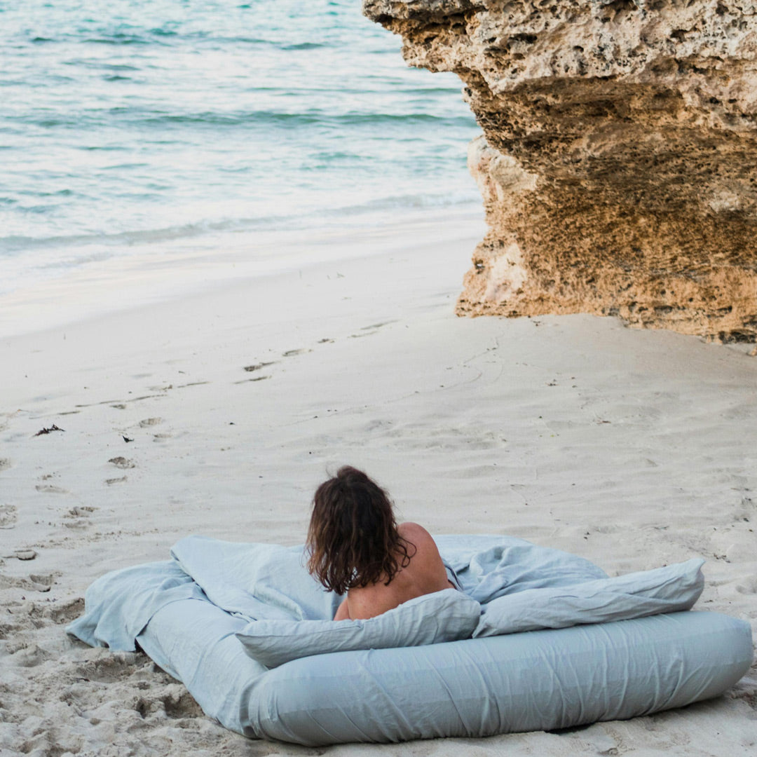 Woman leaning on a blue blanket and mattress by the ocean with cliffs in the background