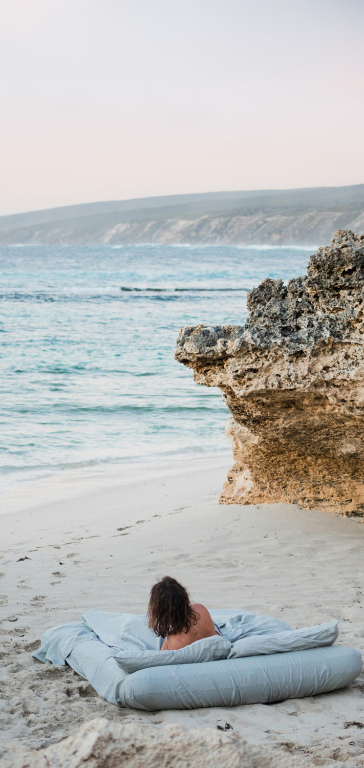 Woman leaning on a blue blanket and mattress by the ocean with cliffs in the background