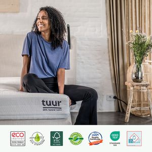 Woman sitting on the Tuur® Original Mattress, with our certificates at the bottom of the picture.
