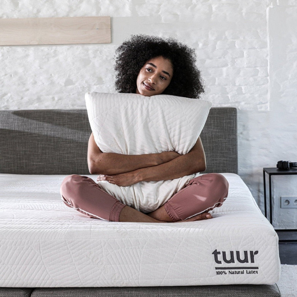 Vrouw die op het Tuur® Spring Matras zit met haar 100% natuurlatex hoofdkussen in de hand.