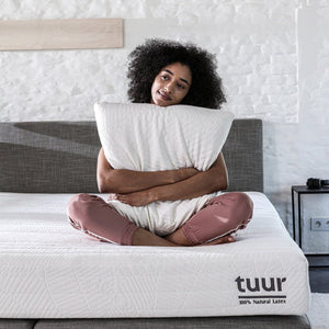 Woman sitting on the Tuur® Spring Mattress with her 100% natural latex pillow in hand.